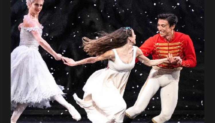 Three people on stage, two women wearing white dresses and the man holding one womens hand wearing a red top and white trousers