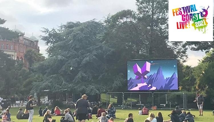 Group of people sitting on the grass in the gardens looking a large screen